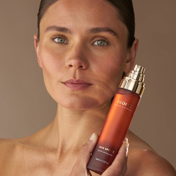 Close-up portrait of a model holding Tropic Sun Spritz facial tanning mist beside her face against a soft neutral background.