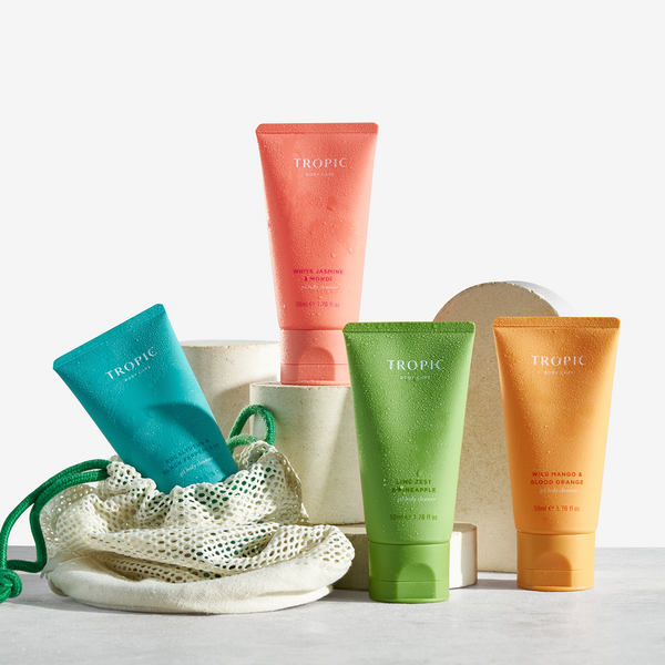 Four Tropic mini gel body cleansers arranged on stone plinths beside a mesh wash bag.