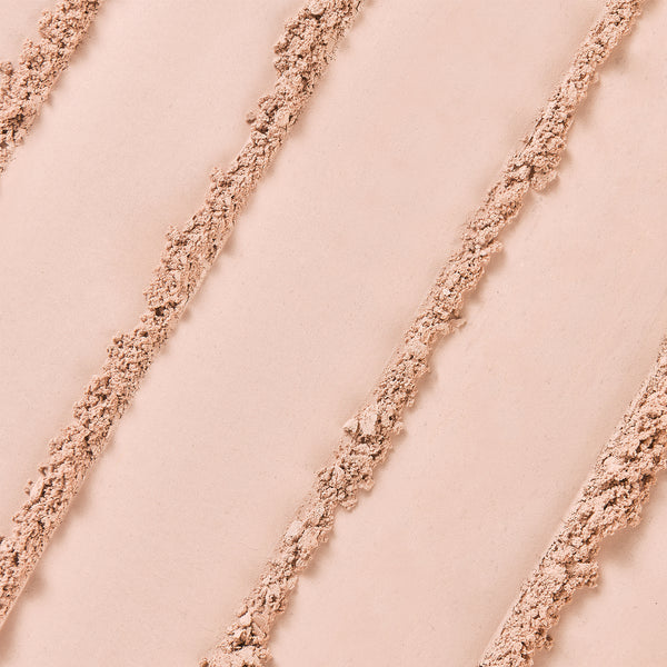 A close-up texture of loose mineral powder foundation in a light beige shade, arranged in diagonal streaks across a smooth surface.