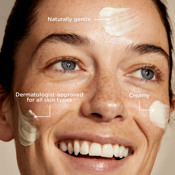 A close-up of a smiling person with light skin and blue eyes, with a few small dabs of smoothing cleanser applied to their face - specifically on the forehead and cheeks. Text annotations on the image highlight that the product is 