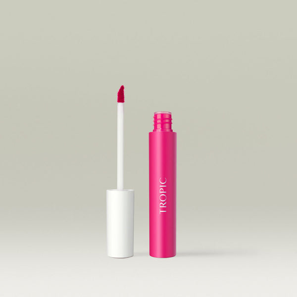 A bright pink liquid lip product with a matching pink tube and white cap, shown open with the applicator wand standing beside it. The angled applicator tip is coated in vibrant pink lip colour against a soft neutral background.