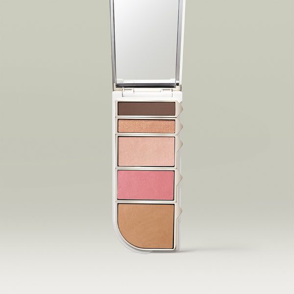 An open makeup palette displayed vertically, featuring five shades: a deep matte brown, a shimmery peach-gold, a soft champagne highlight, a bright pink blush, and a warm bronzer. A mirror is built into the lid above the pans.