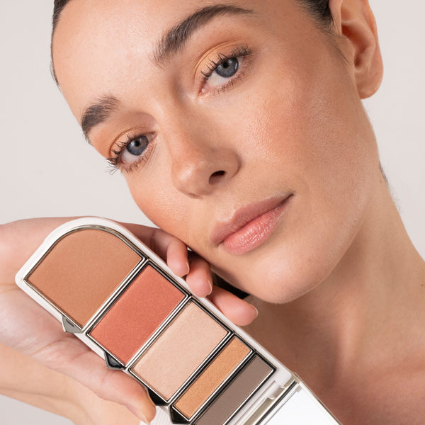 A woman with natural, glowing makeup holds an open makeup palette beside her face. The palette features shades of bronze, coral, champagne, gold, and brown. Her makeup appears to be created using similar warm, neutral tones.