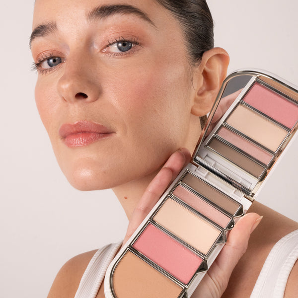 A person with natural makeup holds the Tropic Colour Palette in the “Spring” shade near their face, showing the soft pink, nude, and brown tones applied to their skin.