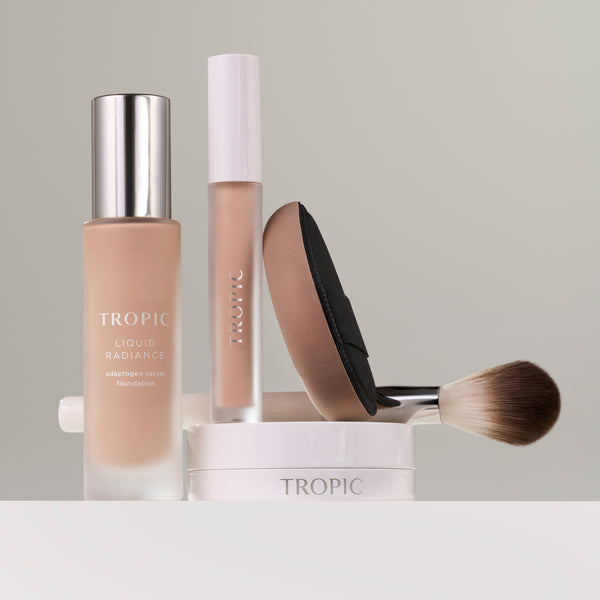 A curated arrangement of Tropic makeup products displayed on a clean, neutral background. The set includes a bottle of Liquid Radiance foundation with a metallic cap, a liquid concealer in a frosted tube, a beige-and-black blending sponge, and a soft, fluffy makeup brush resting on a white Tropic compact.