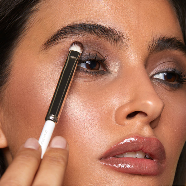 A person applies eyeshadow using a dual-ended makeup brush, blending a soft brown shade on the eyelid for a polished, natural look.