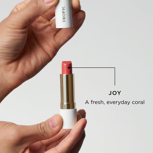 Hands holding an open Tropic lipstick in the shade Joy, described as a fresh everyday coral.