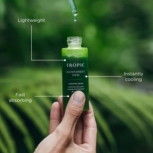 Hand holding an open Tropic Rainforest Dew serum bottle as a dropper releases liquid above it, with text highlighting lightweight, fast-absorbing, and instantly cooling benefits.