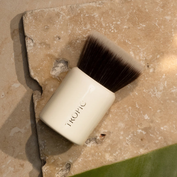 A compact kabuki makeup brush with dense, flat bristles and a glossy cream handle reading “TROPIC,” resting on a textured stone surface in soft, natural lighting.