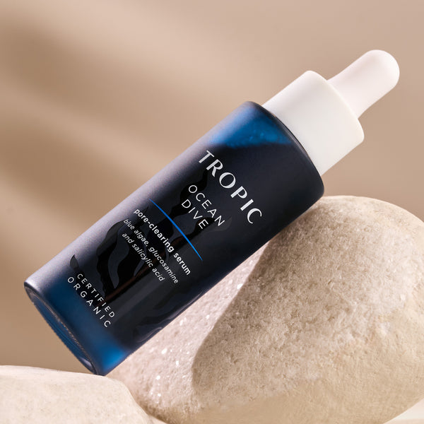 This image showcases a bottle of Ocean Dive, a pore-clearing serum. The dark blue packaging, featuring a dropper applicator, highlights its key ingredients: blue algae, glucosamine and salicylic acid. The product is certified organic and is designed to refine pores, clear skin, and promote a smooth complexion. It is elegantly displayed on a natural stone surface.