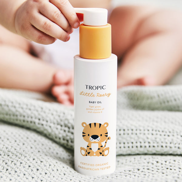 The image showcases a bottle of Tropic Little Roary Baby Oil, held by a baby with soft, cozy blankets in the background. The bottle features a cheerful tiger illustration, appealing to children and parents alike. The label highlights nourishing ingredients such as tiger grass, golden jojoba oil, and vitamin E, which help soothe and moisturise delicate baby skin. The product is marked as Certified Organic and Pediatrician Tested, emphasising its safety and natural formulation.