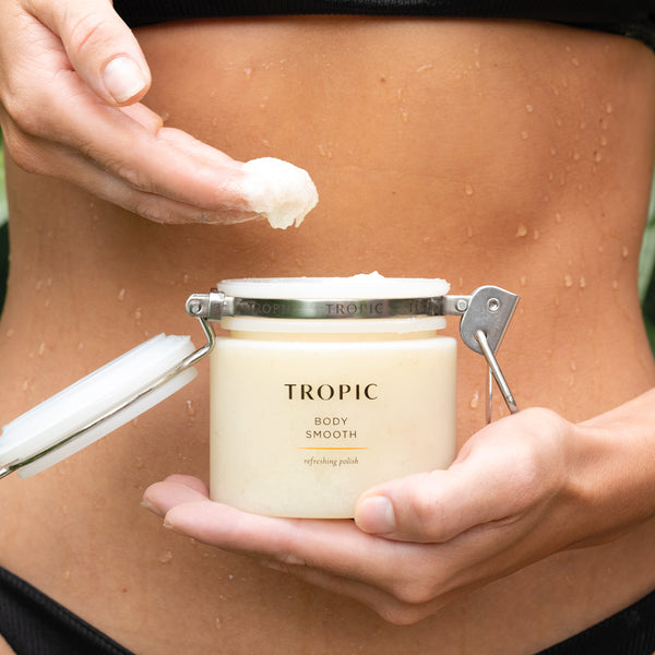 Person holding an open jar of Tropic Body Smooth refreshing polish and applying the creamy scrub to their skin, with water droplets visible on their body.