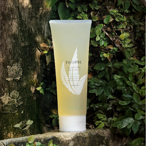 Tropic Clarifying Hair Wash tube placed outdoors against a tree trunk surrounded by lush green foliage, highlighting its natural, plant-powered and eco-friendly formulation.