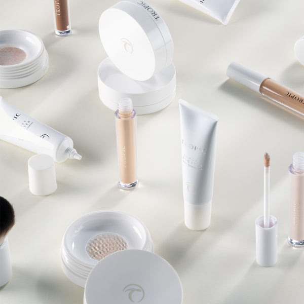A flat-lay arrangement of Tropic base makeup products, including foundation tubes, concealers, cushion compacts, and brushes, all displayed on a soft, light background with gentle shadows for an airy, minimalist aesthetic.