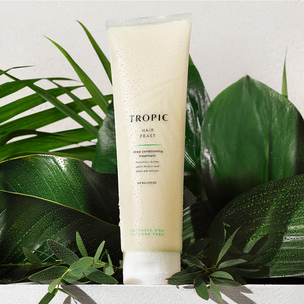 Tropic Hair Feast Deep Conditioning Treatment tube covered in dewy water droplets, surrounded by glossy green tropical leaves, highlighting its nourishing and hydrating formula.