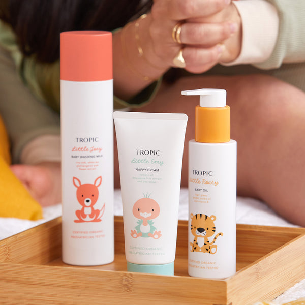 The image features the Tropic Little Heroes Collection, a set of baby care products displayed in a wooden tray. The collection includes:

Little Joey Baby Washing Milk with a kangaroo illustration.
Little Emy Nappy Cream with a bird illustration.
Little Roary Baby Oil with a tiger illustration.
Each product is Certified Organic and Pediatrician Tested, emphasising their gentle and safe formulations for delicate baby skin. The scene captures a warm, nurturing environment, ideal for baby care routines.