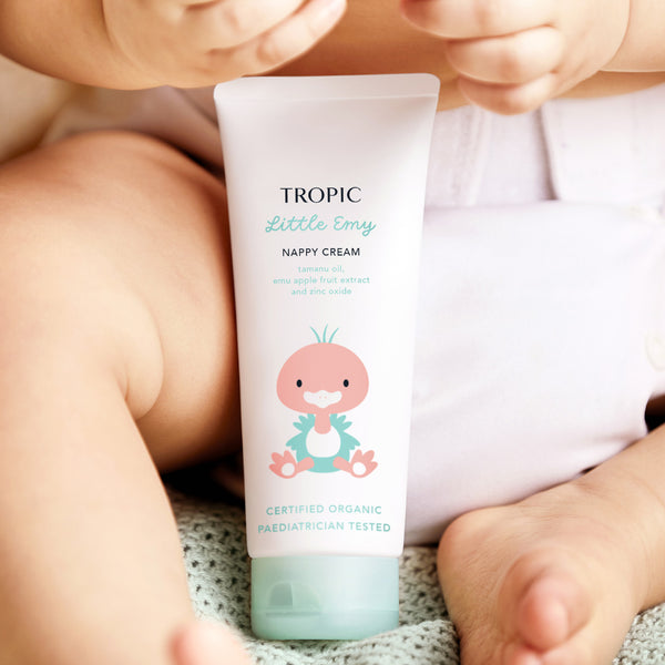 The image displays a tube of Tropic Little Emy Nappy Cream held by a baby, with their legs and diaper visible in the background. The packaging features a cute illustration of a pink bird, creating a playful and child-friendly design. The label highlights key ingredients such as tamanu oil, emu apple fruit extract, and zinc oxide, known for their soothing and protective properties. It is Certified Organic and Pediatrician Tested, emphasising its safety and suitability for delicate baby skin.