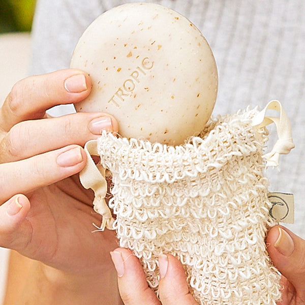 Person holding a round Tropic cleansing body pebble with exfoliating flecks, placing it into a natural cotton mesh pebble pouch.