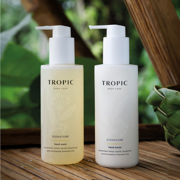 Image shows Tropic Body care Signature hand wash and Signature hand lotion bottles with white  dispensing pumps. There is a faint floral design on the bottle. The products contain Australian lemon myrtle, bergamot and eucalyptus essential oils. The bottles are placed on a wooden surface with green leaves in the background. 