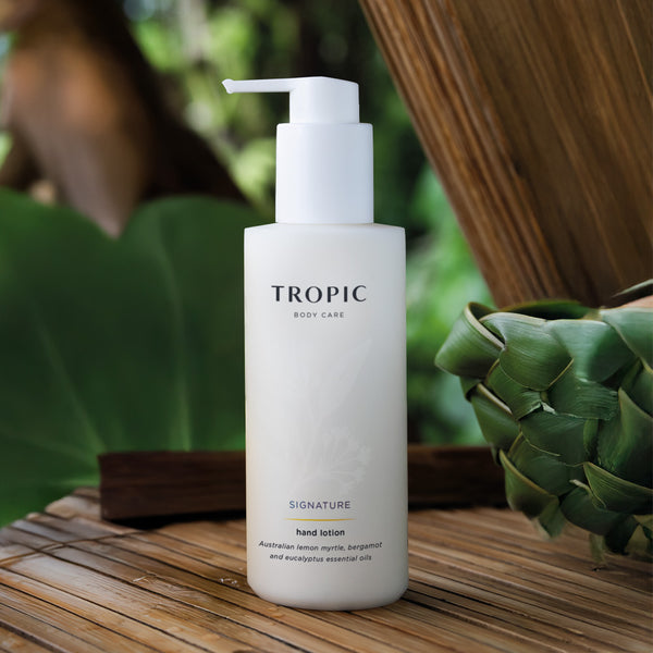 Tropic Signature Hand Lotion bottle displayed in a natural, tropical setting with green foliage and wooden textures, emphasising its botanical and citrus-infused formula.