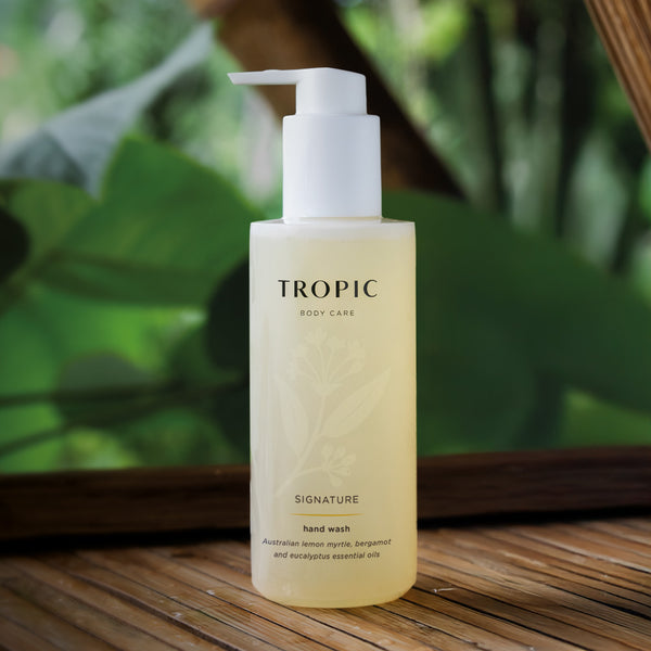 Tropic Signature Hand Wash bottle displayed on a wooden surface with lush green leaves in the background, evoking a natural, botanical freshness.