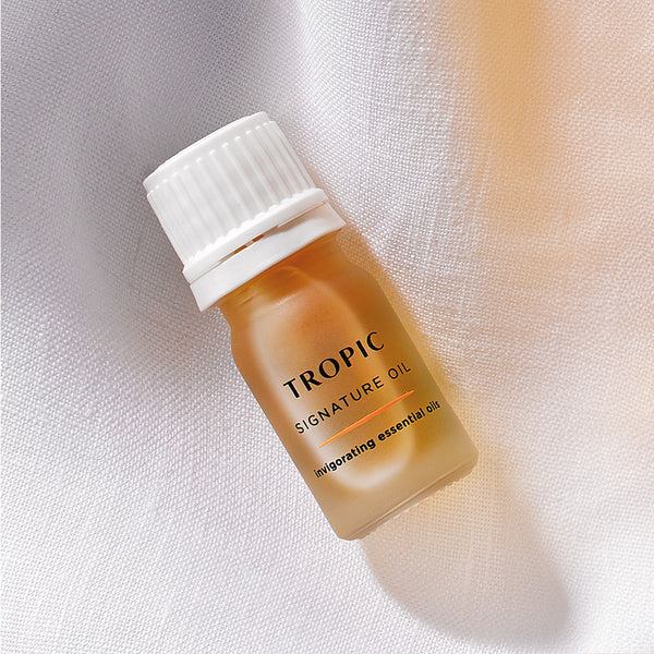 The image features a small frosted bottle of Tropic Signature Oil, placed on a soft, white fabric with warm lighting. The label describes it as containing invigorating essential oils, designed to energize and refresh. 