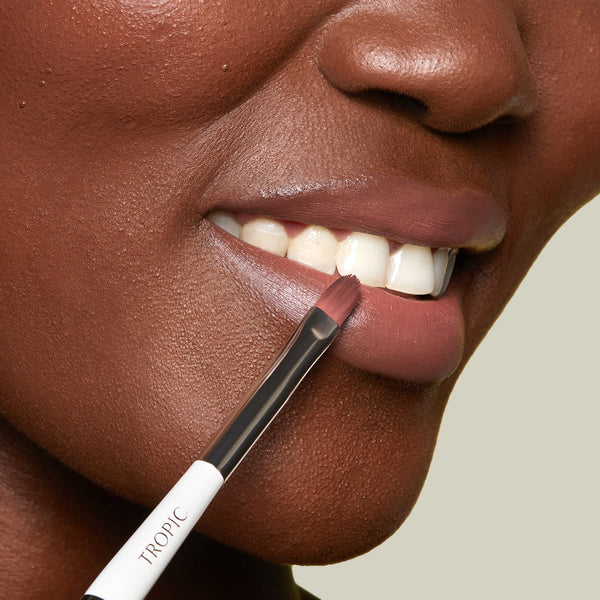 A close-up of a person’s lips as a lip brush applies a smooth, mauve lip colour. The brush, labelled “Tropic,” is gently defining the edge of the lower lip.