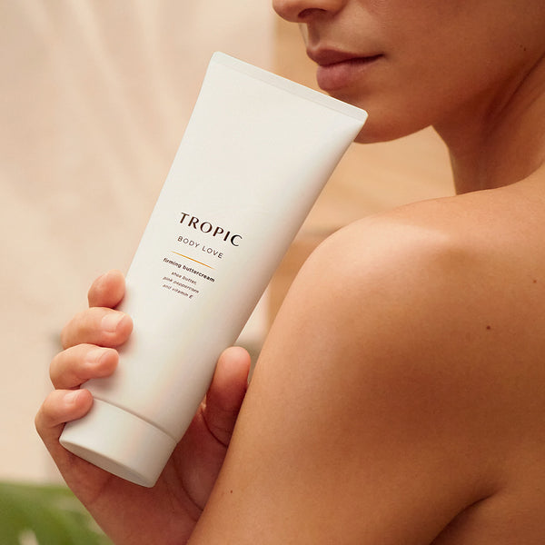 Person holding a white tube of Tropic Body Love firming buttercream near their bare shoulder, with soft lighting highlighting smooth, healthy-looking skin.