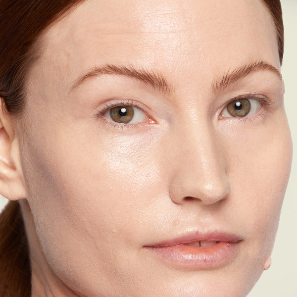 A close-up of a woman with fair skin and light brown hair, showing a dewy, natural-looking complexion with minimal makeup.