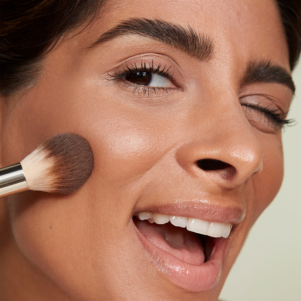 A close-up of a smiling person winking while applying makeup to their cheek with a fluffy powder brush featuring soft brown bristles and a metallic silver ferrule.