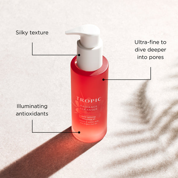A bottle of Tropic Radiance Cleanser with a white pump, set against a light beige background with soft palm leaf shadows. Black text callouts with lines point to the bottle, highlighting its silky texture, ultra-fine pore penetration, and illuminating antioxidants.