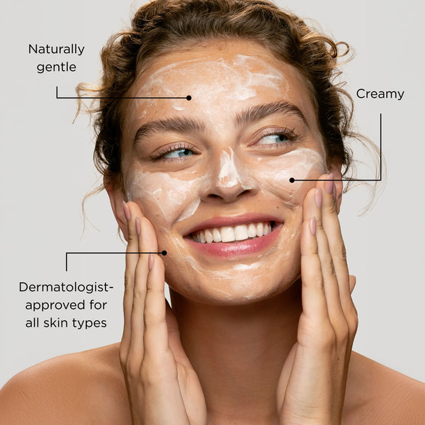 A smiling woman with Smoothing Cleanser spread across her face, with text callouts reading 'Naturally gentle,' 'Creamy,' and 'Dermatologist-approved for all skin types' on a light gray background.