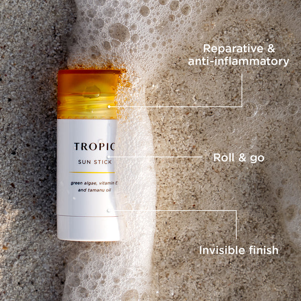 Tropic Sun Stick with a yellow cap lies on wet sand, partially covered by sea foam. Text labels highlight its benefits: “Reparative & anti-inflammatory,” “Roll & go,” and “Invisible finish.”