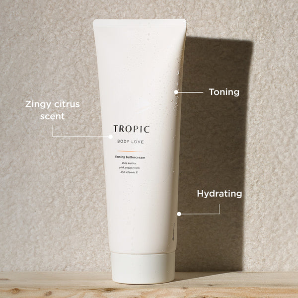 White tube of Tropic Body Love firming buttercream with water droplets on the surface, shown against a textured beige background. Text callouts highlight benefits: “Zingy citrus scent,” “Toning,” and “Hydrating.”
