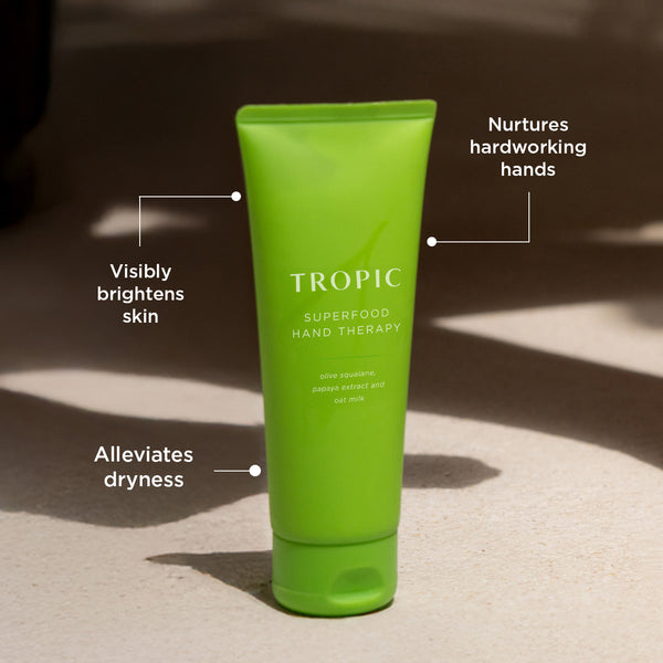 Bright green Tropic Superfood Hand Therapy tube displayed on a sunlit surface with text callouts highlighting benefits: “Visibly brightens skin,” “Alleviates dryness,” and “Nurtures hardworking hands.”