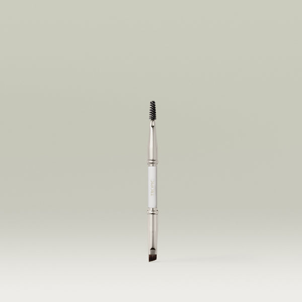 A dual-ended makeup brush featuring a spoolie on one end and an angled brush on the other, shown upright against a soft, neutral background.