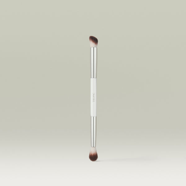 A dual-ended makeup brush with soft, tapered bristles on each end and a sleek white and silver handle, standing upright against a soft neutral background.