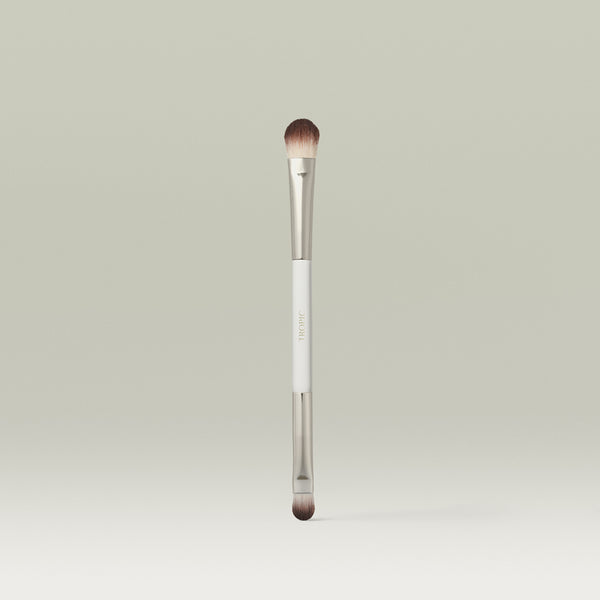 A dual-ended makeup brush with soft, rounded bristles and a sleek metallic finish, standing upright against a soft neutral background.