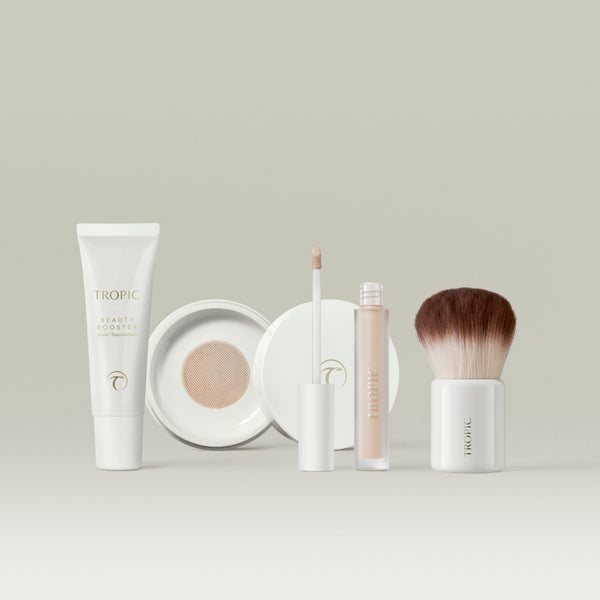 A collection of Tropic base makeup essentials, including a beauty booster foundation tube, a cushion compact with its lid open, a liquid concealer with the wand displayed, and a soft kabuki brush, all arranged neatly against a soft, neutral backdrop. In shade 1.