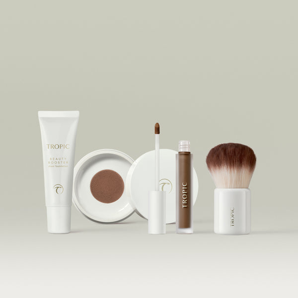 A collection of Tropic base makeup essentials, including a beauty booster foundation tube, a cushion compact with its lid open, a liquid concealer with the wand displayed, and a soft kabuki brush, all arranged neatly against a soft, neutral backdrop. In shade 11.