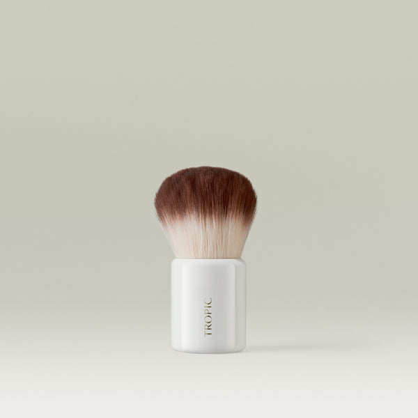 Tropic full-coverage kabuki brush with soft, dense brown-and-white bristles and a glossy white handle, displayed on a light neutral background.