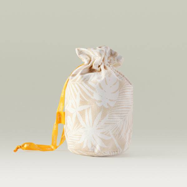 A beige Tropic drawstring gift bag with a white tropical leaf pattern and an orange ribbon featuring the Tropic logo, photographed against a neutral background.