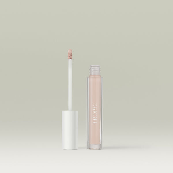 A  liquid concealer in a clear tube labelled “TROPIC,” shown open with its white-capped wand standing beside it against a soft, neutral background. In shade 1.