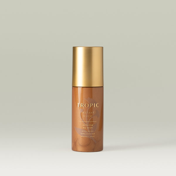Bronze bottle of Tropic Instant Glow perfecting leg serum with a gold cap, containing arnica extract, coffee butter, and vitamins C and E, displayed on a neutral background.