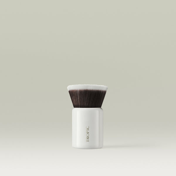 A compact kabuki makeup brush with dense, flat-topped bristles and a glossy white handle, standing upright against a soft, neutral background.