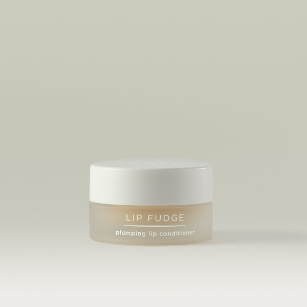 A small frosted clear jar of Lip Fudge plumping lip conditioner with a white lid, shown against a soft, neutral background.