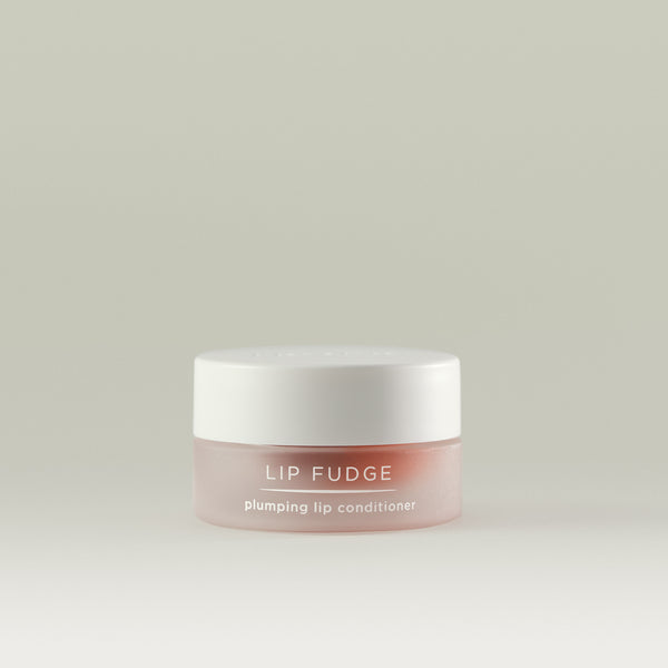 A small frosted rose-toned jar of Lip Fudge plumping lip conditioner with a white lid, shown against a soft, neutral background.