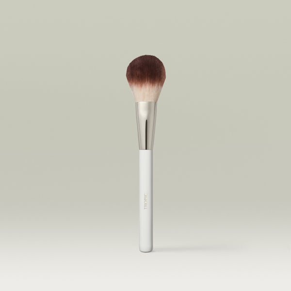 A fluffy, dome-shaped powder brush with soft brown bristles, a metallic silver ferrule, and a white handle, displayed upright against a soft, neutral background.