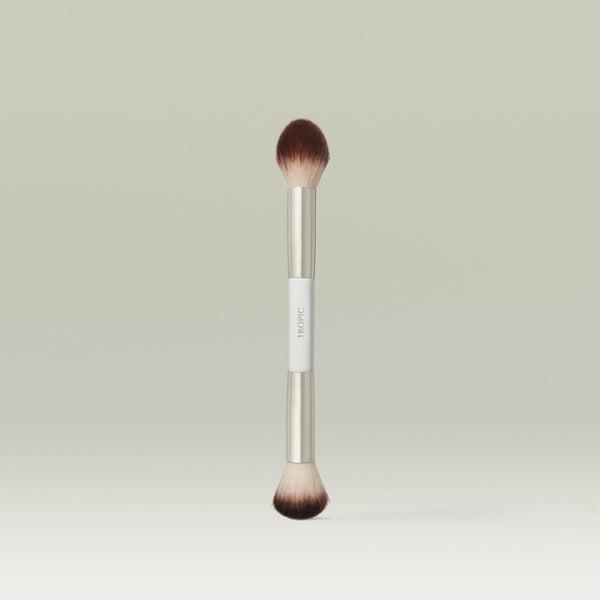 A dual-ended makeup brush with soft, rounded bristles on both ends and a metallic silver ferrule, featuring a white handle section with the brand name TROPI C, shown standing upright against a soft, neutral background.