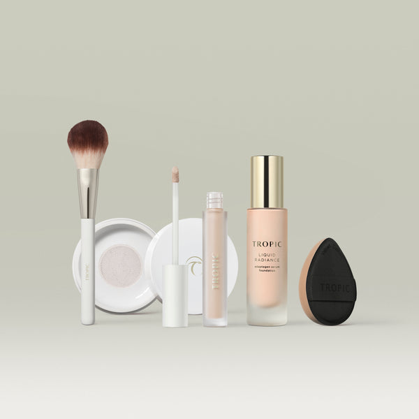 A set of Tropic makeup products in shade 1 arranged in a row against a soft neutral background. The lineup includes a fluffy makeup brush, a white compact, a liquid concealer with a doe-foot applicator, a bottle of Liquid Radiance foundation with a gold cap, and a black teardrop-shaped blending sponge. 
